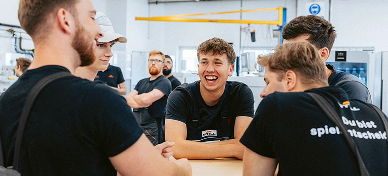 Eine Gruppe junger STILL-Mitarbeiter in schwarzer Arbeitskleidung unterhält sich lachend an einem Tisch in einer Werkstatt.