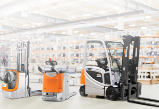 Drei orange-graue STILL-Flurförderzeuge in einer hellen Lagerhalle: ein Hochhubwagen, ein Niederhubwagen und ein Gabelstapler.