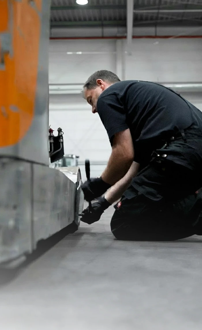 Ein Servicetechniker in schwarzer Arbeitskleidung kniet in einer Lagerhalle auf dem Boden und arbeitet mit einem Werkzeug an einem orange-grauen Flurförderzeug.
