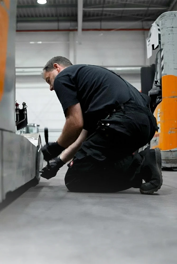 Ein Servicetechniker in schwarzer Arbeitskleidung kniet in einer Lagerhalle auf dem Boden und arbeitet mit einem Werkzeug an einem orange-grauen Flurförderzeug.