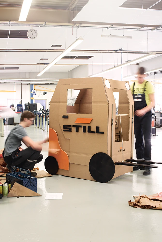 Zwei junge Menschen bauen in einer Werkstatt einen lebensgroßen Gabelstapler mit STILL-Logo aus Karton.