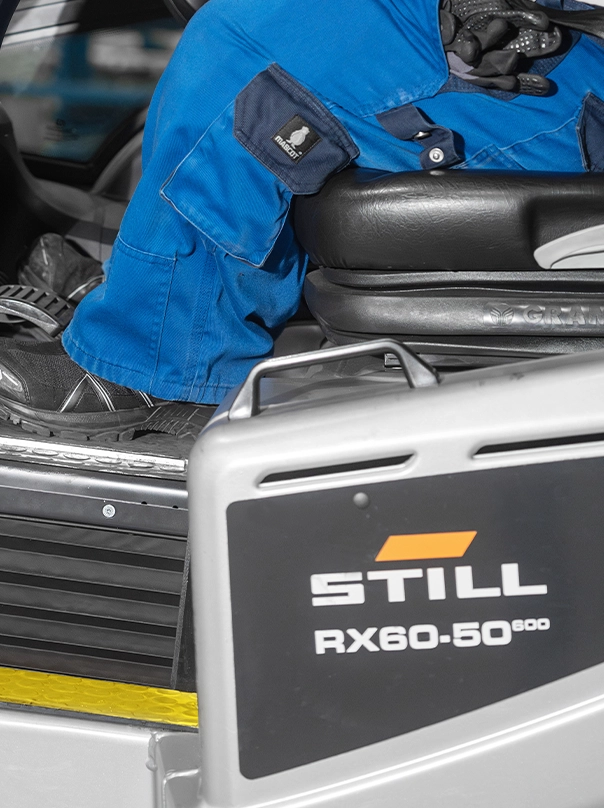 Ein Fahrer in blauer Arbeitskleidung sitzt auf dem Sitz eines STILL RX60-50 Gabelstaplers. Zu sehen sind nur die Beine und die Seite des Staplers mit der Typenbezeichnung.