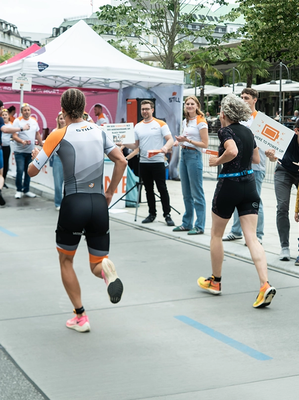 Zwei Triathletinnen laufen an einem STILL-Zelt vorbei, wo Menschen in STILL-Shirts sie mit Schildern anfeuern.