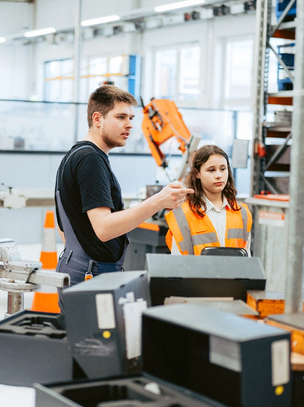 Ein junger Mann in Arbeitslatzhose erklärt einem Mädchen in orangefarbener Warnweste etwas in einer Produktionshalle.