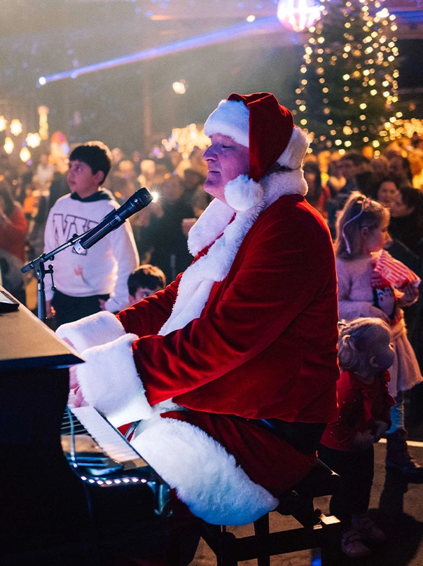 Ein als Weihnachtsmann verkleideter Mann spielt bei einer Weihnachtsveranstaltung Klavier und singt, im Hintergrund stehen Kinder und ein beleuchteter Weihnachtsbaum.