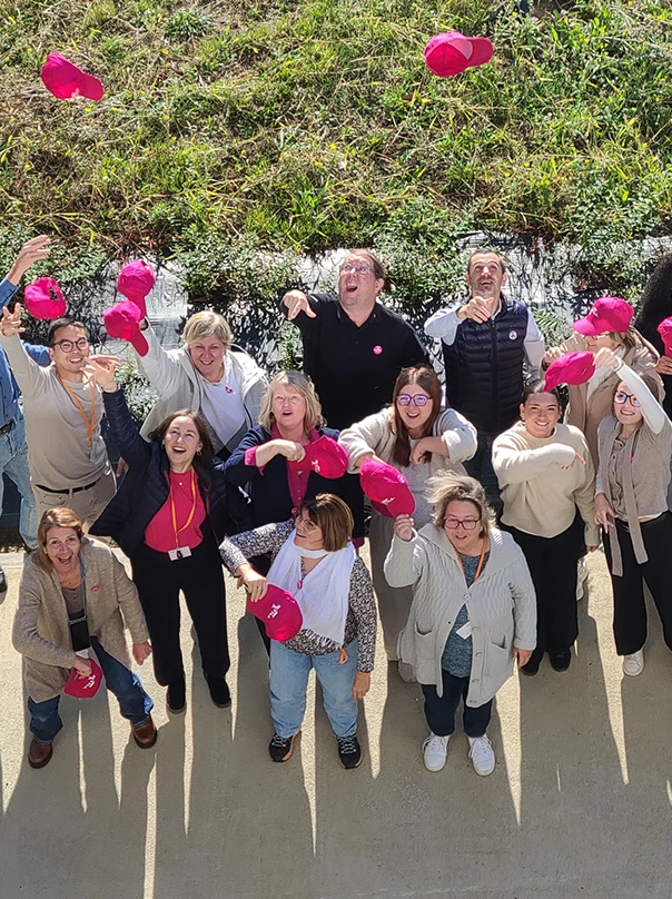 Eine fröhliche Gruppe von Mitarbeitenden wirft pinke Kappen in die Luft, aufgenommen von oben.