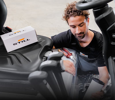 Ein STILL-Servicetechniker arbeitet mit einer Filterpatrone in der Hand im Motorraum eines Gabelstaplers, auf dem Sitz liegt ein STILL-Ersatzteilkarton.