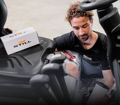 Ein STILL-Servicetechniker arbeitet mit einer Filterpatrone in der Hand im Motorraum eines Gabelstaplers, auf dem Sitz liegt ein STILL-Ersatzteilkarton.