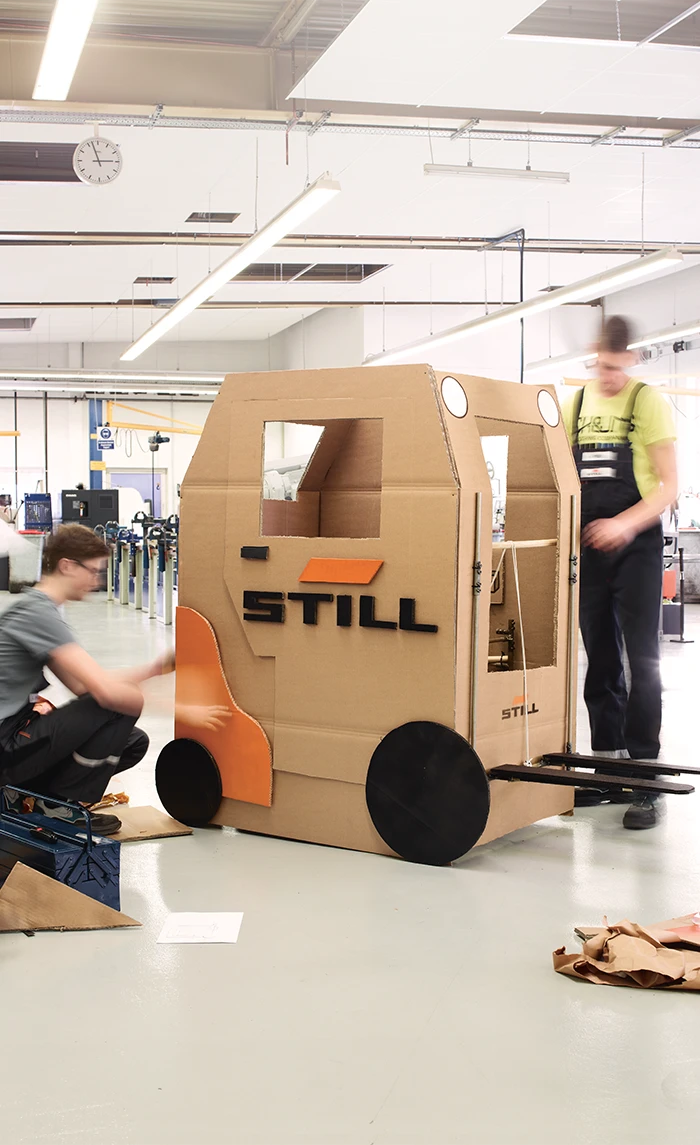 Two young people build a life-size forklift out of cardboard with the STILL logo in a workshop.