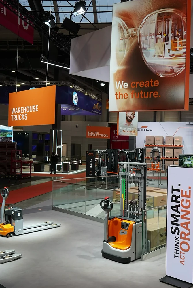 STILL trade fair stand with orange and grey industrial trucks, a “Warehouse Trucks” sign, and banners reading “We create the future.” and “Think smart. Act orange.”