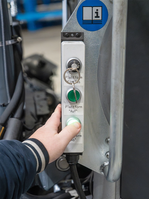 A hand presses an illuminated button on a control panel for a platform on an industrial truck. The panel has a key switch and buttons labelled “Plattform Ein/Aus” (platform on/off), “Plattform Auf” (platform up) and “Plattform Ab” (platform down).