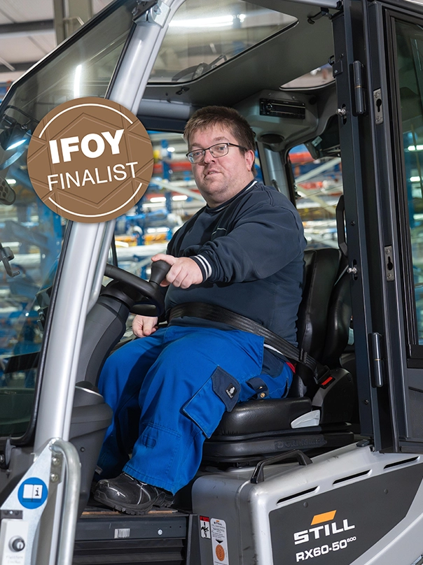 A man with glasses, wearing a seat belt, sits at the wheel of a STILL RX60-50 forklift, with an “IFOY Finalist” badge.