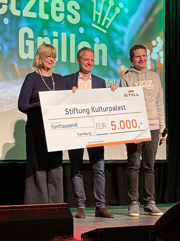 Three people on a stage hold up a large STILL donation cheque for €5,000 made out to the Stiftung Kulturpalast in Hamburg.