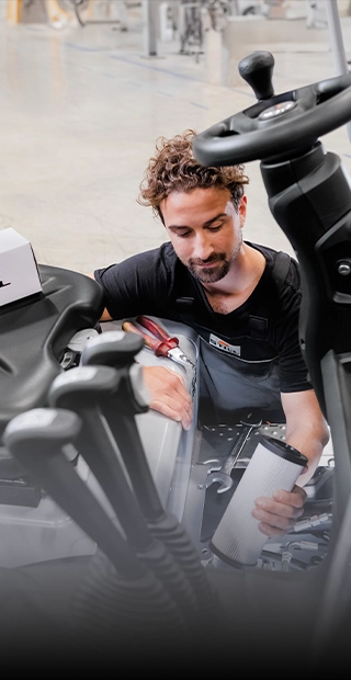 A STILL service technician with a filter cartridge in his hand works in the engine compartment of a forklift, with a STILL spare parts box on the seat.