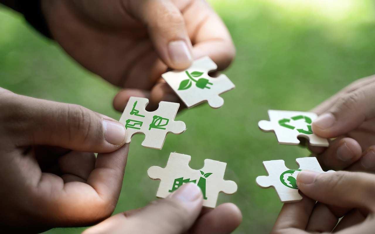 Several hands hold puzzle pieces with green sustainability symbols, such as a recycling sign, a plug with leaves and a globe, and bring them together.